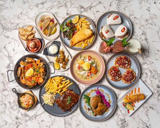 A table spread of SUGO'S signature dishes and cocktails — tacos, fish and chips, paella, lamb and more on a marble table