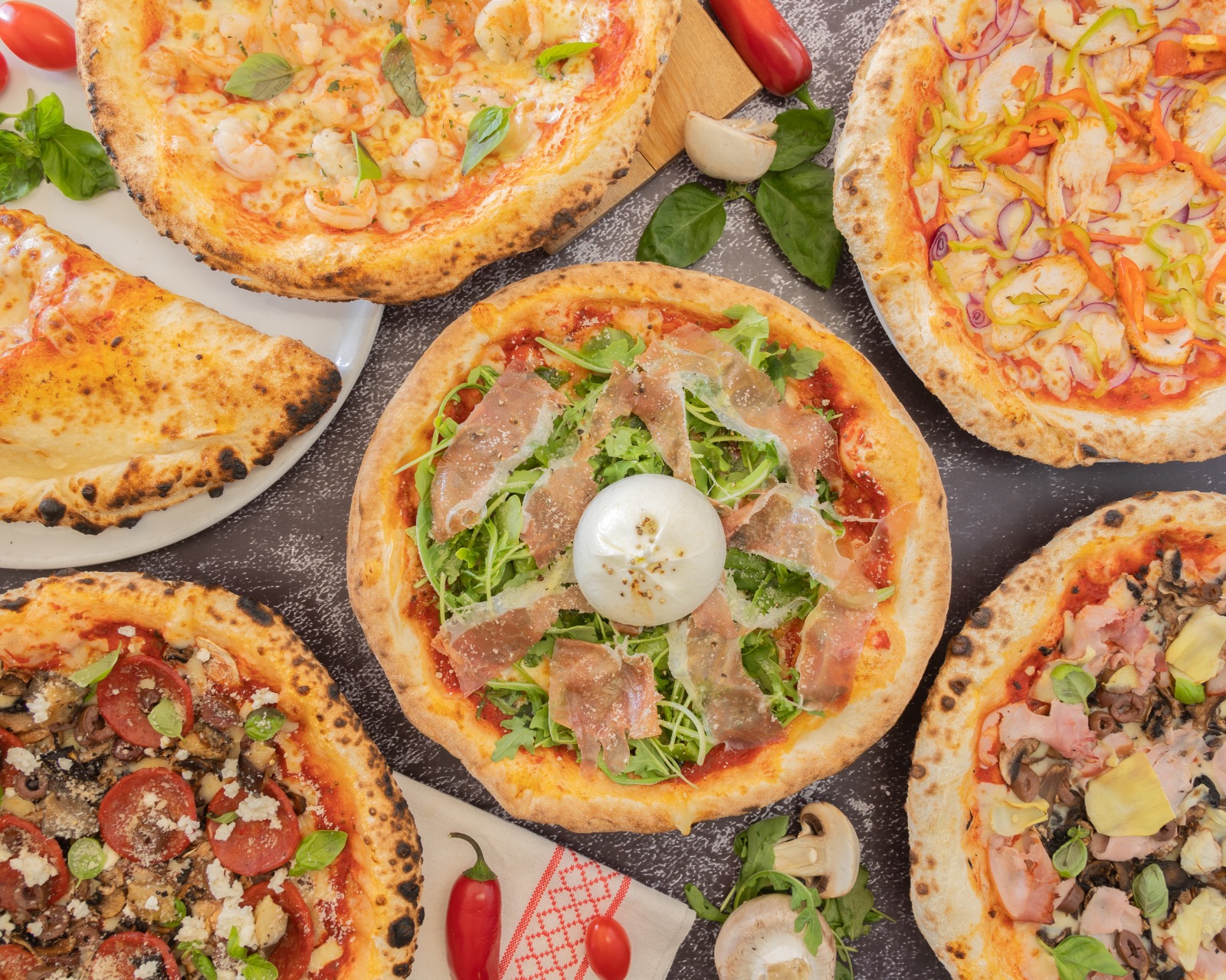 A spread of wood-fired pizzas with prosciutto, rocket, salami, mushrooms and roasted chicken on a dark table